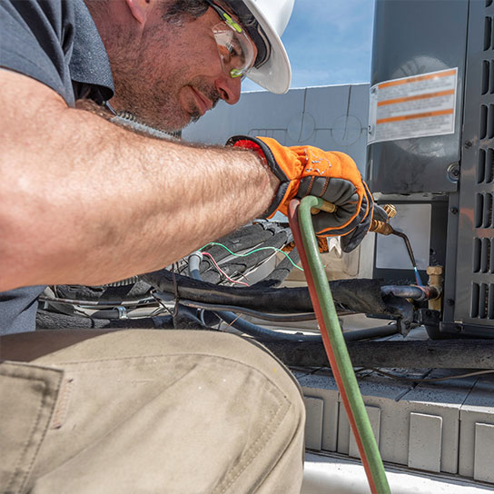 comm-maint-tech-544x544-v1 An HVAC technician on the roof of a business repairing a commercial air conditioner